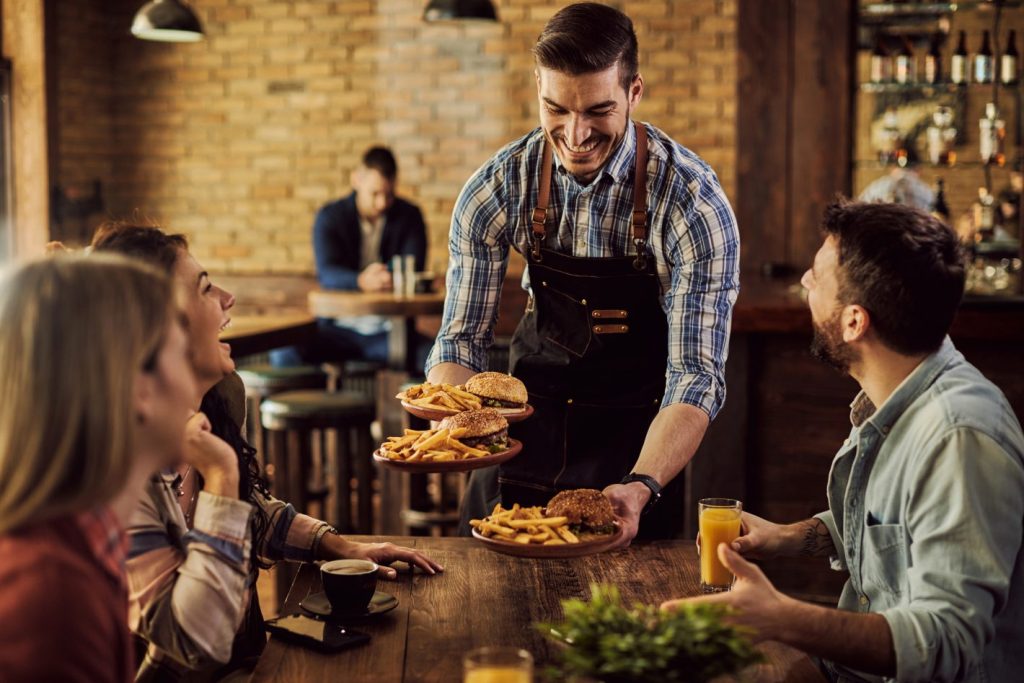 Promoções de Black Friday no seu restaurante: aproveite | Foto de um garçom servindo hambúrgueres em uma mesa com clientes felizes | Atacadão