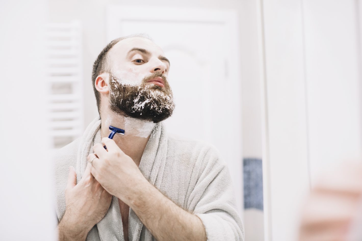 Produtos para tirar barba: conheça os melhores | Homem tirando a barba com lâmina se olhando no espelho | Atacadão