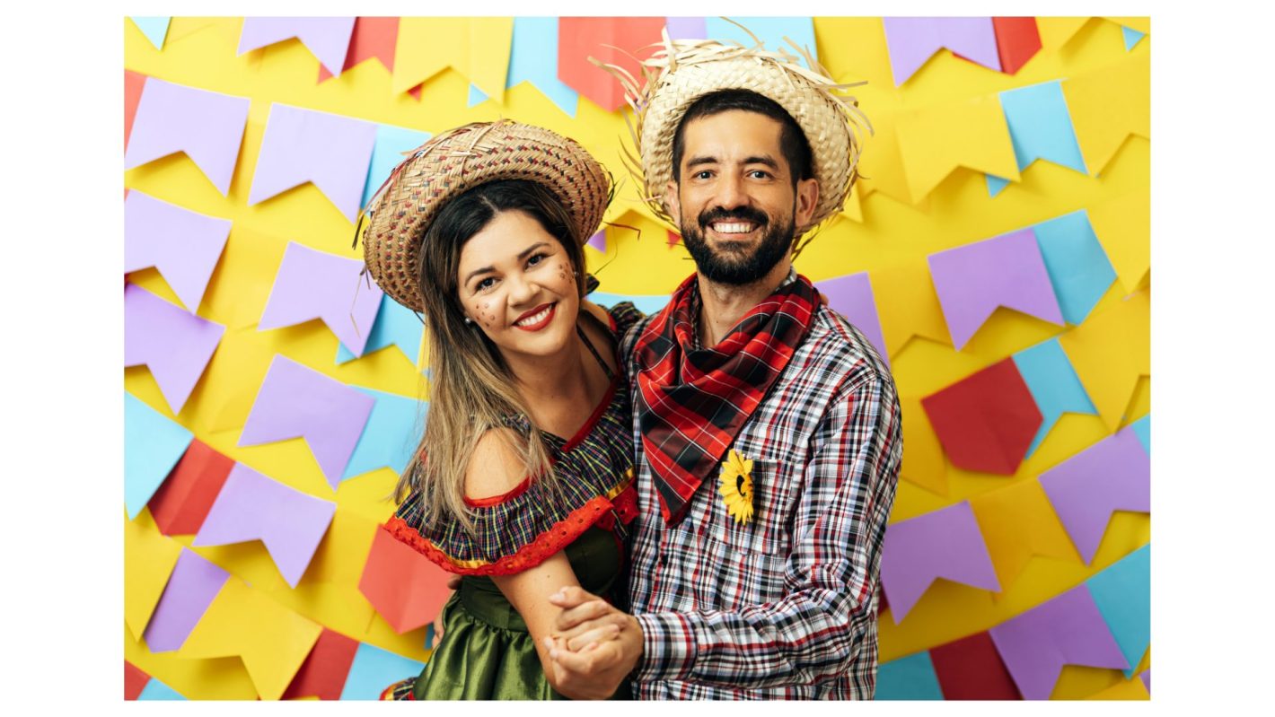 Dicas de festa junina: como celebrar em casa? | Foto de um casal caracterizados com roupas de festa junina em frente a bandeirinhas coloridas.| Atacadão
