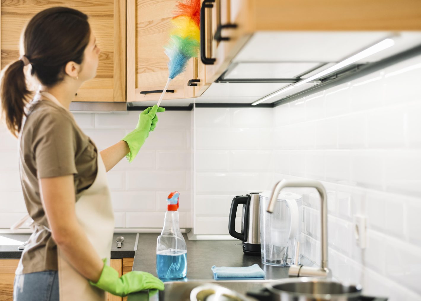 Como fazer uma limpeza pesada na cozinha | Foto da vista lateral de uma mulher usando luvas verdes de borracha, ela segura um espanador. | Atacadão