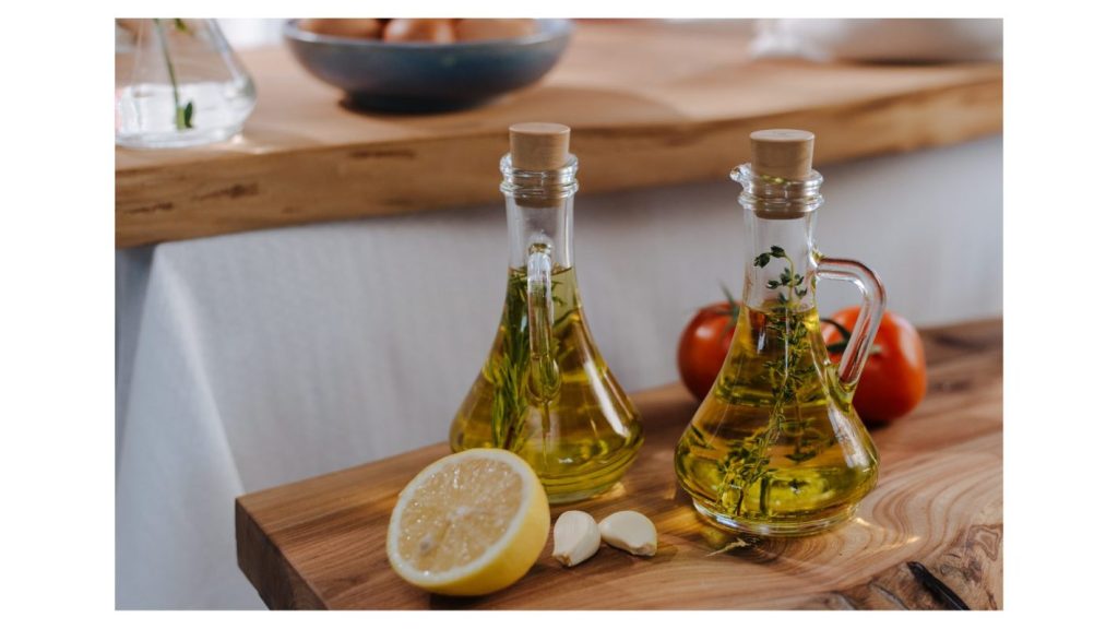 As vantagens de utilizar azeite vão além de um sabor incrível | Foto de dois galheteiros, contendo azeites aromatizados com alecrim, sobre uma mesa de madeira. | Atacadão